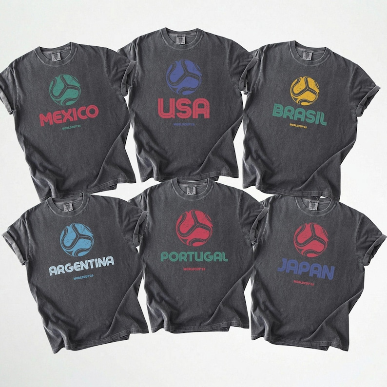 May include: Six dark grey t-shirts with country names and soccer ball graphics. The shirts feature the names Mexico, USA, Brasil, Argentina, Portugal, and Japan in various colours. Each shirt has a distressed, vintage look.