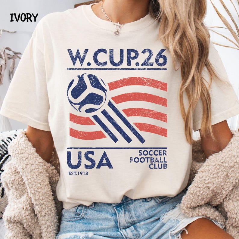 A model wearing an ivory cream t-shirt with a navy and red distressed USA W.CUP.26 soccer club design, paired with a casual cardigan.