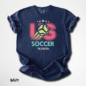 Flat lay of a navy blue t-shirt featuring a bold US Soccer W.CUP.26 graphic with pink and turquoise elements.