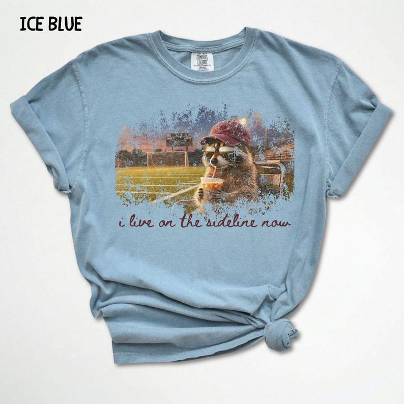 Pode incluir: Camiseta azul gelo com um guaxinim usando chap&eacute;u e &oacute;culos de sol, bebendo de um copo, com o texto "i live on the sideline now". O fundo mostra um campo esportivo.