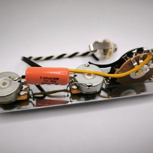Op de afbeelding: Een chromen gitaarbedieningsplaat met drie potentiometers, een condensator en een schakelaar. De oranje condensator heeft zwarte tekst. De draden zijn geel, zwart en wit. Een gitaarjack is ook inbegrepen.