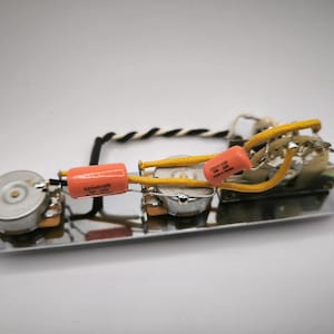 Może przedstawiać: Płytka kontrolna gitary z elementami elektronicznymi. Płytka jest srebrna z dwoma pomarańczowymi kondensatorami, żółtym okablowaniem i czarno-białym skręconym przewodem. Elementy są zamontowane na płytce.