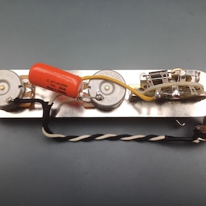 Op de afbeelding: Gitaarelektronica-assemblage met drie potentiometers, een oranje condensator en een schakelaar, allemaal gemonteerd op een metalen plaat. Zwarte, witte en gele draden verbinden de componenten. Aan het einde is een jack te zien.