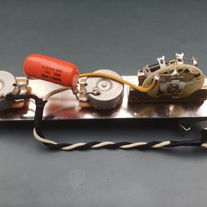 Pode incluir: Conjunto eletrónico de guitarra com potenciómetros, um condensador e um interruptor montados numa placa metálica. O condensador laranja tem o texto "0.047uF 600V TAO 2210 Made in Mexico". Fios e uma tomada também são visíveis.