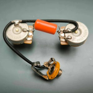 Puede incluir: Arnés de cableado de guitarra con dos potenciómetros, un condensador naranja y un cable negro. El arnés incluye un conector para conectar a un amplificador. Los componentes están dispuestos sobre una superficie gris.