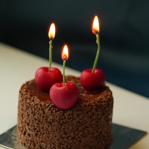 May include: A small chocolate cake covered in chocolate flakes, adorned with three cherry-shaped candles with lit flames. The cake rests on a silver square plate, against a dark blue backdrop.