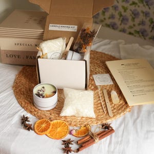 May include: A candle-making kit featuring a cardboard box, a white tin candle, and various ingredients. The kit includes wax, dried orange slices, star anise, cinnamon sticks, and instructions. The box is labelled "Candle Making Kit."