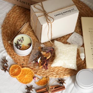 May include: A candle-making kit with a white box tied with twine, a candle tin, and ingredients like dried orange slices, star anise, and wax. The kit includes the text "Candle Making Kit" and "the joy is in the making."