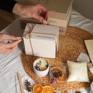 May include: A candle making kit with a white box tied with twine, a candle tin, dried orange slices, star anise, and a bag of wax beads. The kit includes the text "CANDLE MAKING KIT".