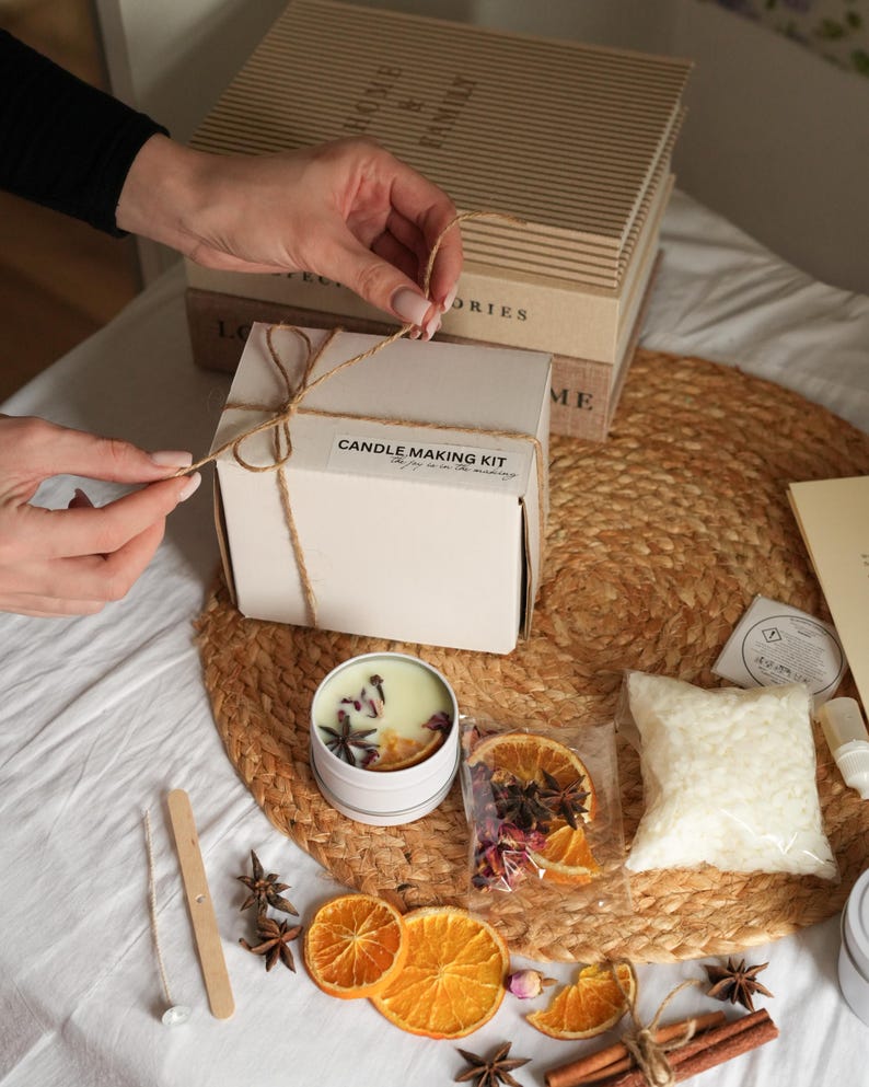 May include: A candle making kit with a white box tied with twine, a candle tin, dried orange slices, star anise, and a bag of wax beads. The kit includes the text "CANDLE MAKING KIT".