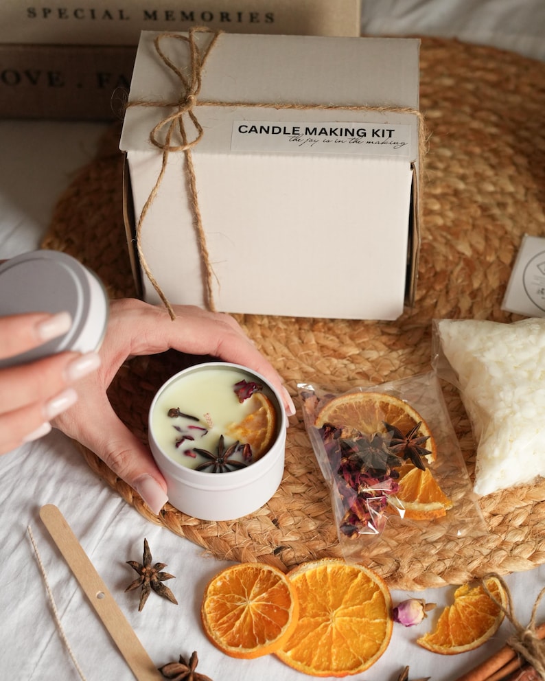 May include: A candle making kit with a white box tied with twine. The kit includes a white candle tin with dried orange slices, star anise, and rose petals. The text on the box reads "Candle Making Kit".