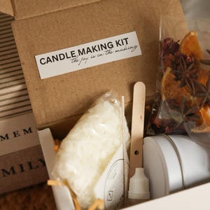 May include: A candle making kit in a brown cardboard box. The kit includes a bag of wax, a wooden stirring stick, a bottle, and a white container. A label on the box reads "CANDLE MAKING KIT the joy is in the making".