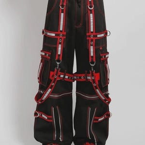 Könnte beinhalten: Schwarze Cargohose mit roten Akzenten, einschließlich Taschen, silbernen Reißverschlüssen und reflektierenden Streifen. Die Hose mit weitem Bein ist mit D-Ringen und Riemen verziert. Rote Nähte und ein kantiger, moderner Stil definieren das Design.