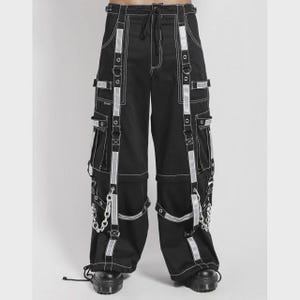 May include: Black cargo pants with white stitching and reflective silver accents. The pants feature multiple pockets, straps, and silver chain details. The pants have a drawstring waist and are worn with black boots.
