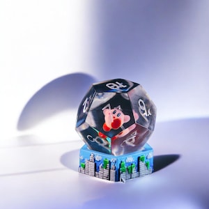 May include: A clear, multi-sided dice with numbers 1 through 9 printed on each side. The dice is sitting on a small, blue and green cityscape base. A pink and red character is visible inside the dice.