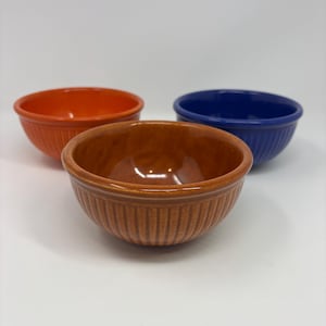 May include: Three small, round ceramic bowls with a ribbed exterior texture. One bowl is bright orange, one is deep blue, and the front-most bowl is a warm, earthy brown. Each bowl features a smooth, glossy finish and a simple, open design.