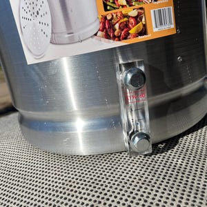 May include: A silver aluminum pot with a clear plastic sight glass with red markings. The sight glass has two silver bolts on either end. The pot has a silver lid with holes in it. The pot is sitting on a gray mesh surface.