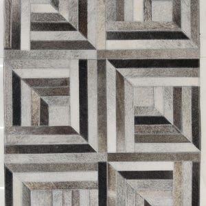 May include: A rectangular rug with a geometric pattern. The rug features a patchwork design with squares and rectangles in shades of gray, black, and white. The rug is made of a soft, textured material, likely animal hide.