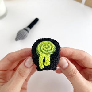 May include: A small, black, crocheted cauldron featuring a bright lime green, swirled, textured liquid spilling over the rim. The item is crafted from yarn and displays a rounded shape with a flat base, showcasing intricate stitching throughout.