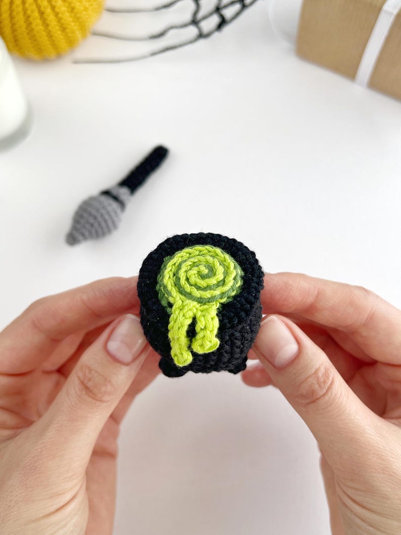 May include: A small, black, crocheted cauldron featuring a bright lime green, swirled, textured liquid spilling over the rim. The item is crafted from yarn and displays a rounded shape with a flat base, showcasing intricate stitching throughout.
