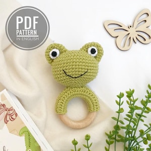 Könnte beinhalten: Eine gehäkelte grüne Frosch-Rassel mit einem Holzring. Der Frosch hat schwarze Augen und einen lächelnden Mund. Die Rassel liegt auf einem weißen Hintergrund.