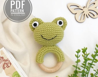 Op de afbeelding: Een gehaakte groene kikker rammelaar met een houten ring. De kikker heeft zwarte ogen en een glimlachende mond. De rammelaar ligt op een witte achtergrond.