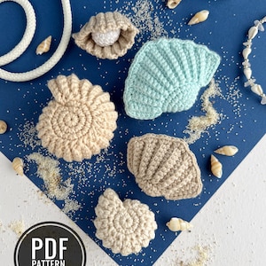 Può includere: Una collezione di cinque conchiglie all'uncinetto in beige e azzurro, tra cui una conchiglia a spirale e una vongola con una perla bianca. Gli oggetti sono disposti con piccole conchiglie naturali e corda bianca. Un badge circolare riporta PDF PATTERN IN ENGLISH.