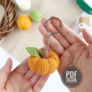 Peut inclure: Un porte-clés en forme de citrouille crochetée, aux couleurs orange, verte et marron. La citrouille a une surface texturée et est attachée à un porte-clés argenté. Le porte-clés est tenu dans les mains d'une personne. Le texte "PDF PATTERN IN ENGLISH" est visible.
