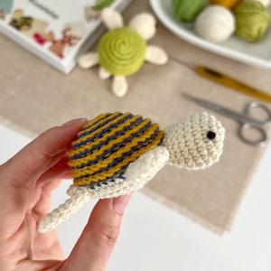 Puede incluir: Una tortuga de ganchillo hecha a mano con un caparaz&oacute;n a rayas amarillas y azules, cabeza color crema y peque&ntilde;os ojos negros. La tortuga se sostiene en una mano, con otros suministros de artesan&iacute;a en el fondo, incluyendo hilo y un libro.