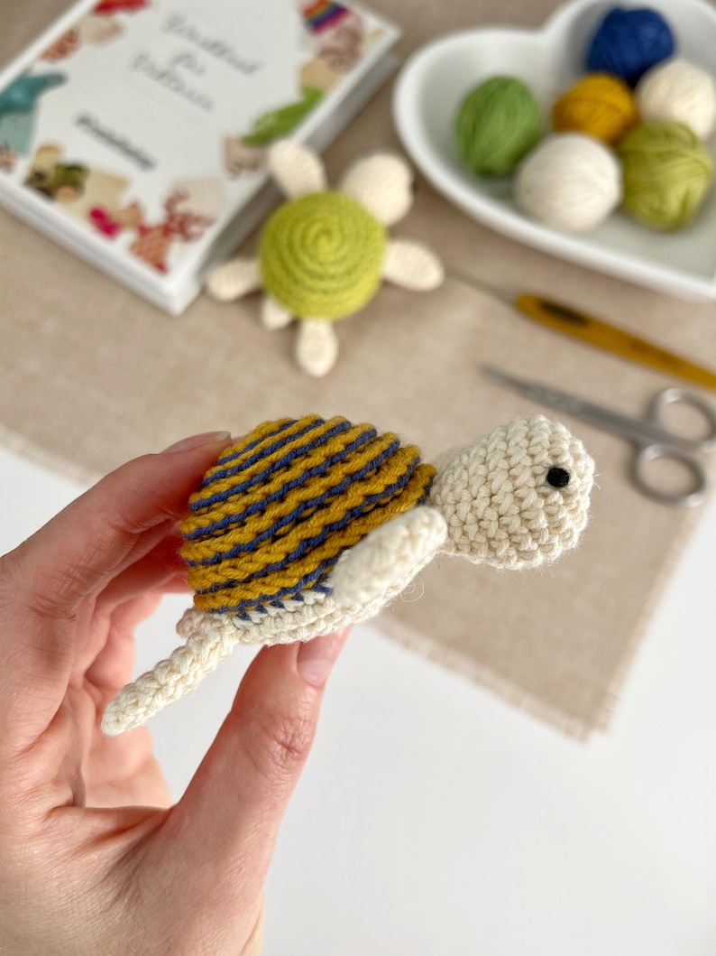 Puede incluir: Una tortuga de ganchillo hecha a mano con un caparaz&oacute;n a rayas amarillas y azules, cabeza color crema y peque&ntilde;os ojos negros. La tortuga se sostiene en una mano, con otros suministros de artesan&iacute;a en el fondo, incluyendo hilo y un libro.