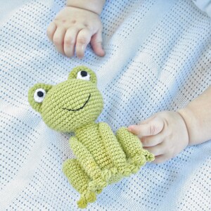 Op de afbeelding: Een lichtgroen gehaakt kikker speelgoed met zwart-witte ogen en een lachende mond. De kikker ligt op een lichtblauwe deken. Het speelgoed wordt vastgehouden door een baby.