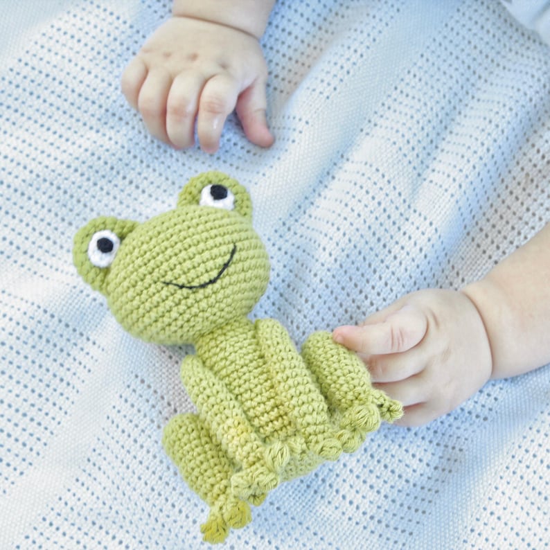 Op de afbeelding: Een lichtgroen gehaakt kikker speelgoed met zwart-witte ogen en een lachende mond. De kikker ligt op een lichtblauwe deken. Het speelgoed wordt vastgehouden door een baby.
