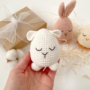 May include: Three crocheted Easter egg-shaped stuffed animals: a white lamb, a pink bunny, and a yellow chick. Each animal has a closed-eye face with embroidered details.