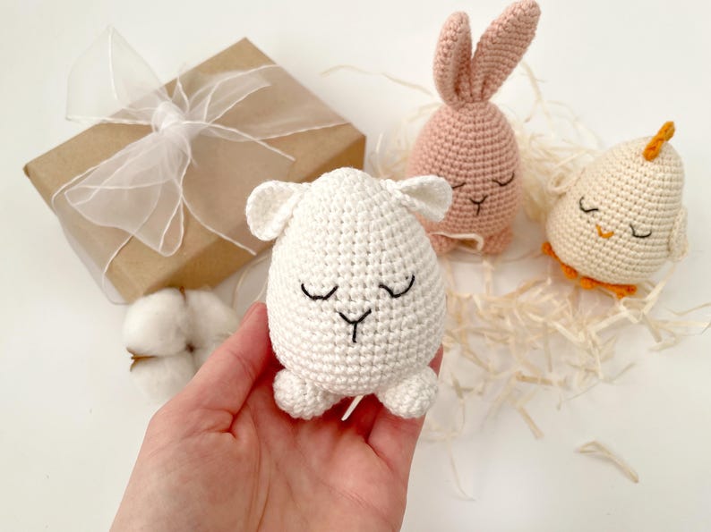 May include: Three crocheted Easter egg-shaped stuffed animals: a white lamb, a pink bunny, and a yellow chick. Each animal has a closed-eye face with embroidered details.