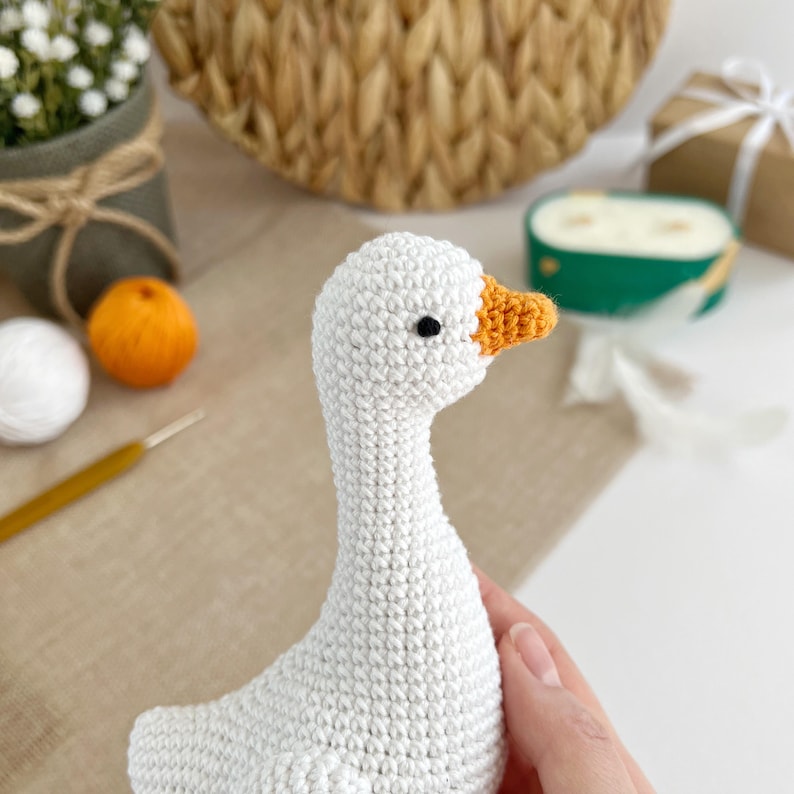 May include: A handmade white crochet goose with an orange beak and black eyes. The goose is a soft, plush toy, perfect for nursery decor or as a unique gift. The crochet stitches are visible, adding to the handcrafted charm.