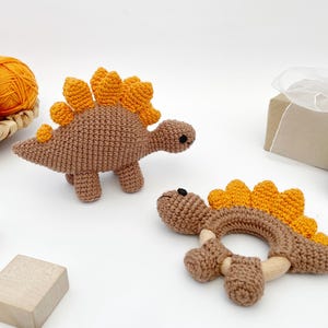 May include: Two crochet dinosaur toys, one standing and one a teething ring. The dinosaurs are brown with orange spines. A basket of yarn and a wrapped gift are in the background. Wooden blocks are in the foreground.
