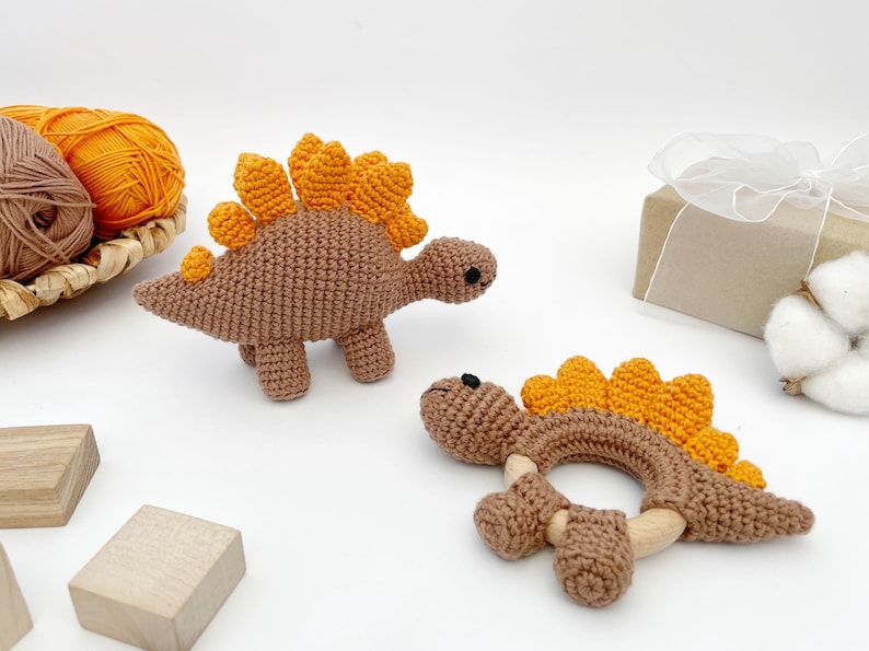 May include: Two crochet dinosaur toys, one standing and one a teething ring. The dinosaurs are brown with orange spines. A basket of yarn and a wrapped gift are in the background. Wooden blocks are in the foreground.