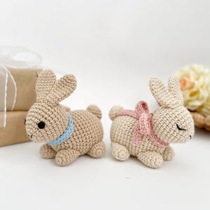 May include: Two crocheted bunny rabbits, one with a blue ribbon and the other with a pink ribbon. The bunnies are sitting on a white surface.