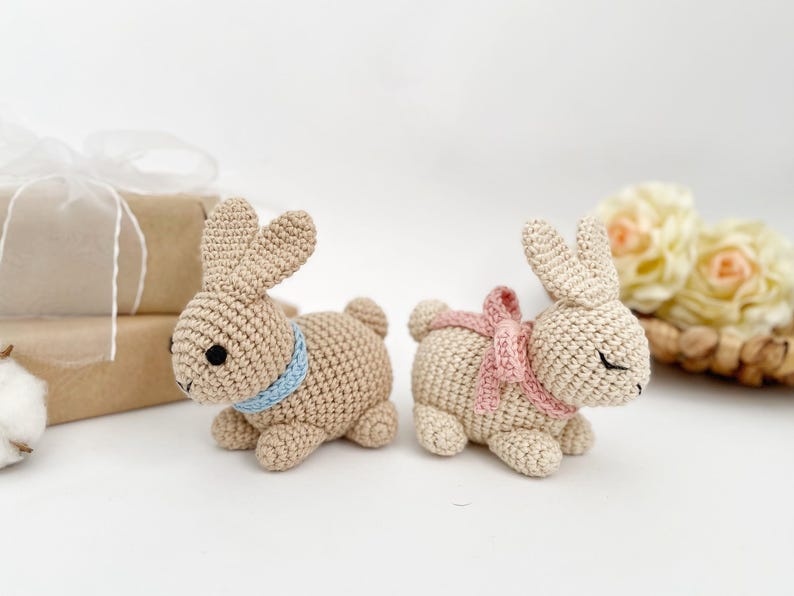 May include: Two crocheted bunny rabbits, one with a blue ribbon and the other with a pink ribbon. The bunnies are sitting on a white surface.