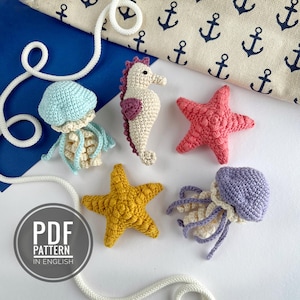 May include: A collection of handmade crochet sea creatures, including a seahorse, starfish, and jellyfish. The seahorse is off-white with a purple accent. The starfish are pink and yellow. The jellyfish are blue and purple. A PDF pattern in English is visible.