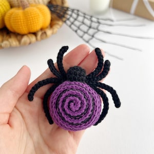 May include: A crocheted spider ornament featuring a round body with a purple and black spiral pattern. The spider has a small black head and eight black crocheted legs extending from the sides. The yarn texture is visible throughout the piece.