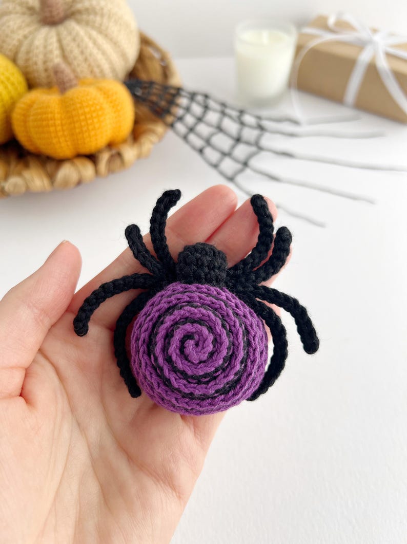 May include: A crocheted spider ornament featuring a round body with a purple and black spiral pattern. The spider has a small black head and eight black crocheted legs extending from the sides. The yarn texture is visible throughout the piece.