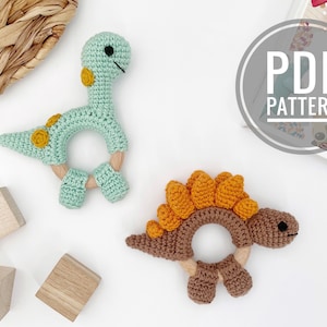 Op de afbeelding: Twee gehaakte dinosaurus-bijtringen, een groene met gele accenten en een bruine met oranje accenten. Beide bijtringen hebben houten ringen in het midden.