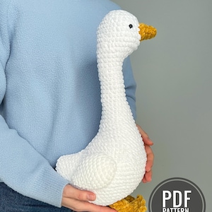 May include: A white crochet goose with a yellow beak and feet. The goose is held by a person wearing a light blue jumper and jeans. The image includes a circular graphic that says "PDF PATTERN IN ENGLISH".