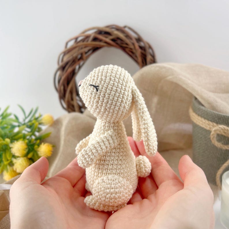 May include: A crocheted cream-colored bunny rabbit with black eyes and a pink nose. The bunny is sitting in the palm of a person's hand.
