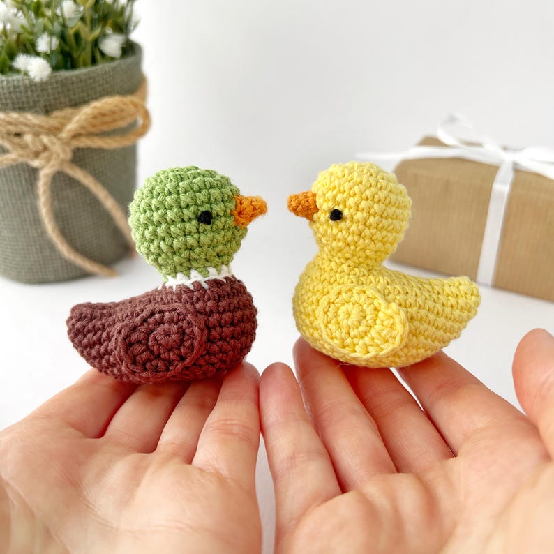 May include: Two handmade crochet ducklings. One duckling is yellow with an orange beak, and the other is brown with a green head and orange beak. The ducklings are held in open hands.