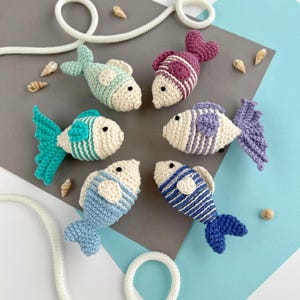 May include: A collection of six crocheted fish in various colors, including teal, purple, and blue, with white accents. The fish are arranged on a gray and blue surface, with seashells and a white rope adding to the nautical theme. Each fish has a unique striped or patterned design.