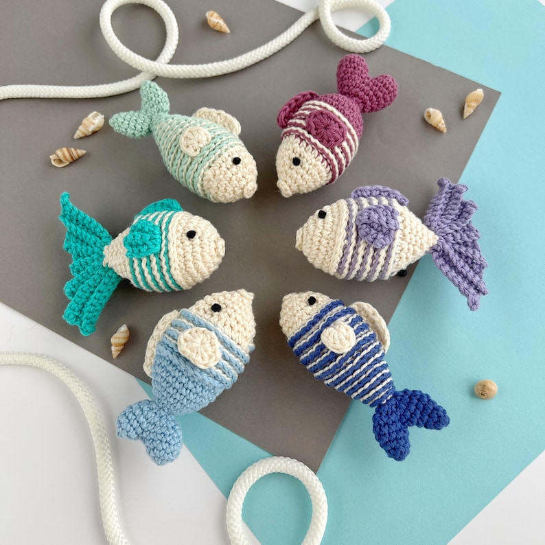 May include: A collection of six crocheted fish in various colors, including teal, purple, and blue, with white accents. The fish are arranged on a gray and blue surface, with seashells and a white rope adding to the nautical theme. Each fish has a unique striped or patterned design.