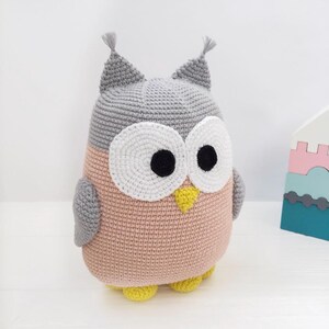 May include: A crocheted owl plush toy with gray and pink yarn. The owl has large white eyes with black pupils and a yellow beak. The owl is sitting on a white surface.