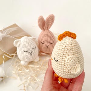 May include: Three crocheted stuffed animals shaped like an egg. The animals are a pink bunny, a white chick, and a white lamb. The animals have embroidered eyes and mouths.
